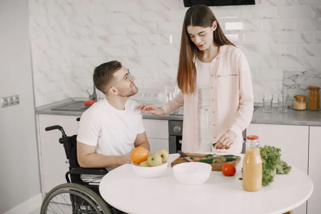 A man using a wheelchair and a woman preparing fresh vegetables together in a bright kitchen