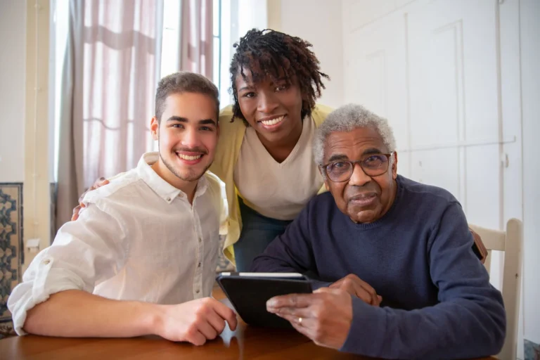 Supervised living support: two smiling support staff, a Black woman and a young man, sit with an older Black man holding a tablet at a kitchen table